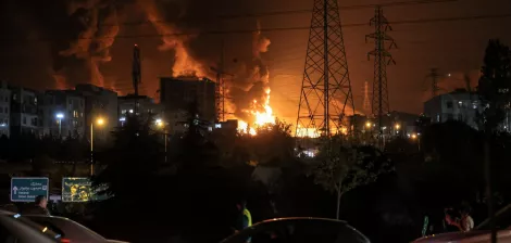 En la imagen, hay un gran incendio en una ciudad nocturna, con humo y luces. Autos circulan en primer plano.