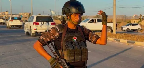 Un homme armé, en tenue militaire, se tient sur une route avec des voitures en arrière-plan.