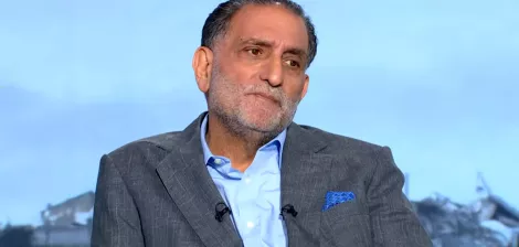 A middle-aged man in a suit with a serious expression, seated against a blurred background.