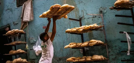 Un hombre levanta canastas con pan en un ambiente de panadería. Las paredes son azules y desgastadas.