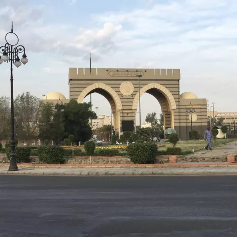 A large archway with decorative towers, surrounded by greenery and a road.