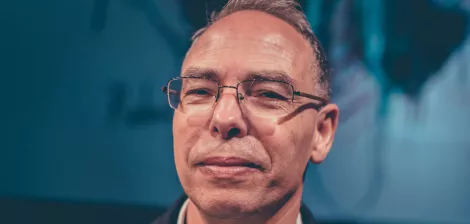 A middle-aged man with glasses and a slight smile, standing against a blurred background.