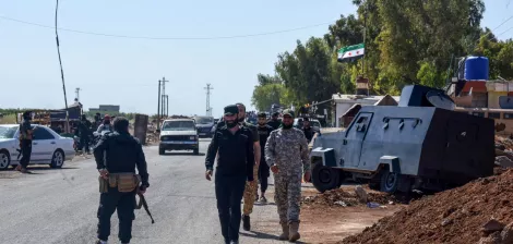 تصویر، یک خیابان با خودروها و کامیون‌ها است. چند نفر با لباس نظامی در حال حرکت هستند. دیوارها و درختان اطراف هستند.