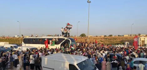 A large crowd gathers, waving flags and celebrating, with vehicles parked around.