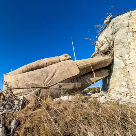 The image shows a large, partially fallen stone statue lying on its side against a rocky background, surrounded by sparse vegetation and a clear blue sky.