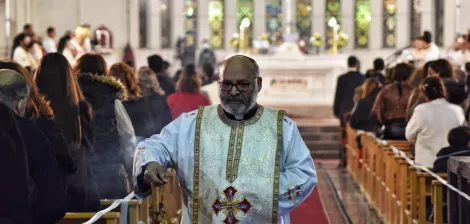 A priest in white robes walks down the aisle of a church during a service.