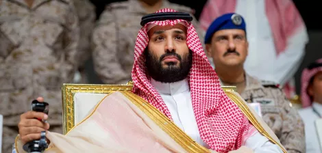 A man in traditional Saudi attire sits on a throne-like chair, looking serious and dignified.