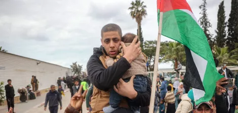 The image depicts a scene of a gathering or demonstration. A man is holding a child closely to him, showcasing a protective gesture. Around them, a crowd appears to be rallying, with some individuals raising their hands and a prominent Palestinian flag visible in the background. The atmosphere seems charged, indicating a moment of significance, possibly related to a political or social issue. The setting includes trees and a building in the background, against a cloudy sky.