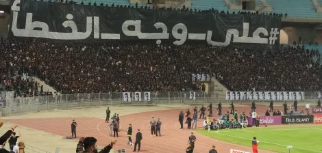 A large crowd at a stadium with a black banner and fans cheering passionately.