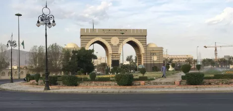A grand archway with domes, surrounded by greenery and a road, under a cloudy sky.