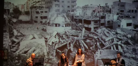 Four performers sit with instruments in front of a large backdrop of war-torn buildings.