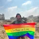 Un soldat souriant tient un drapeau arc-en-ciel sur un terrain dévasté.