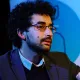 Homme avec des lunettes, cheveux bouclés, en discussion, vêtu d'une veste.
