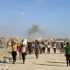 Des personnes marchent dans des décombres, portant des colis, avec de la fumée au loin.