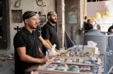 Trois hommes à un stand distribuant des sandwichs, en arrière-plan des bâtiments.