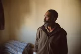 Un homme avec une barbe, regard pensif, se tient dans une pièce simple.