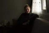 Femme souriante, assise sur un canapé, lumière douce filtrée par un rideau.