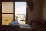 Chambre simple avec lit, fenêtre ouverte sur un paysage urbain et ciel nuageux.