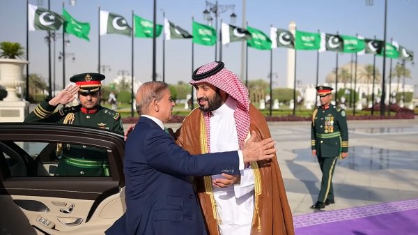 Deux hommes en costume échangent une accolade, avec des drapeaux en arrière-plan.