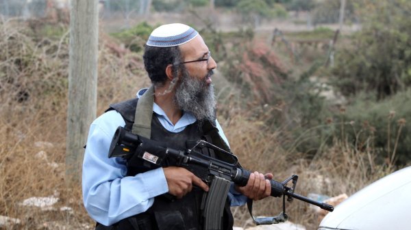 L'image montre un homme qui porte une kippa et une veste de protection. Il est armé d'un fusil automatique et se tient près d'une voiture dans un environnement extérieur, avec de la végétation et des débris autour de lui. Son expression et sa posture suggèrent qu'il est en alerte ou surveille les alentours.
