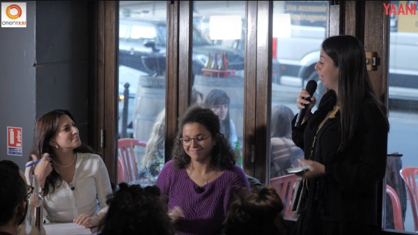 L'image montre une scène intérieure où plusieurs personnes sont assises à des tables. Une femme se tient debout au premier plan, tenant un microphone, probablement en train de parler ou de présenter quelque chose. Elle porte des vêtements noirs et semble engagée dans une conversation dynamique. À sa gauche, une autre femme sourit, tandis qu'à sa droite, une femme avec des lunettes apporte une expression de concentration. À l'arrière-plan, on peut voir des clients assis, créant une atmosphère animée. Les grandes fenêtres laissent entrer la lumière et montrent une vue partielle de l'extérieur.