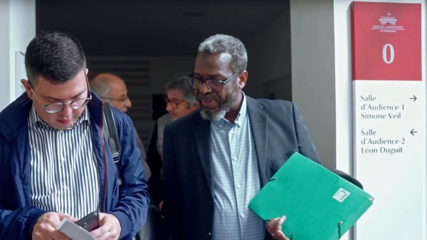 L'image montre un groupe de personnes dans un couloir apparemment nimbé d'une ambiance professionnelle. Deux hommes au premier plan interagissent, l'un tenant un téléphone et l'autre un dossier vert. L'environnement semble être celui d'un bâtiment public ou d'un tribunal, avec des panneaux indiquant les salles d'audience. Les visages des personnes sont engagés dans une conversation, ce qui suggère un échange collaboratif ou une discussion importante.