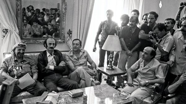 Réunion de personnages politiques dans une salle, nombreux spectateurs en arrière-plan.