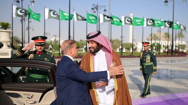 Deux hommes en costume échangent une accolade, avec des drapeaux en arrière-plan.