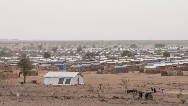 L'image montre un large camp de tentes dans une zone aride. On y voit de nombreuses petites tentes de couleur bleue et beige, entourées de sols secs et caillouteux, avec quelques arbres dispersés dans le paysage.