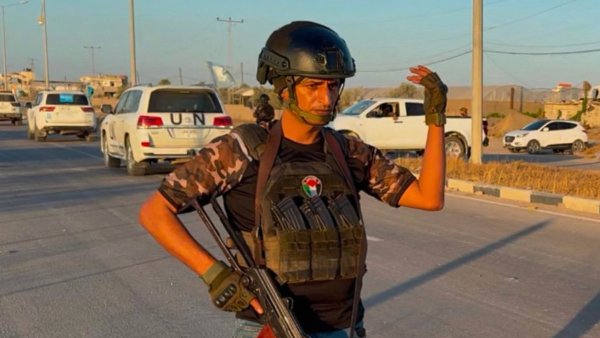 Un homme armé, en tenue militaire, se tient sur une route avec des voitures en arrière-plan.