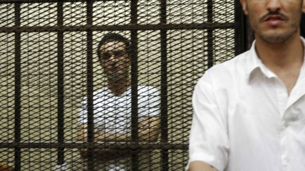 Deux hommes se tiennent derrière des barreaux, l'un visible et l'autre en arrière-plan.
