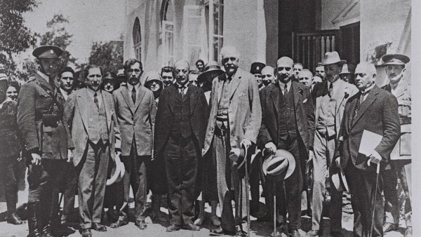 Un groupe d'hommes en costumes, certains avec des chapeaux, posant ensemble à l'extérieur.