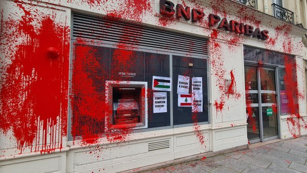 L'image montre une agence de la BNP Paribas, dont la façade est recouverte de peinture rouge. Des affiches sont visibles sur la vitrine, et l'automate de distribution de billets est également marqué par cette peinture. L'ensemble donne une impression de vandalisme ou de protestation, soulignant peut-être un message politique ou social.