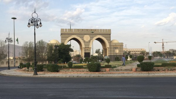 Un grand portail avec arches, entouré de verdure et de lampadaires, sous un ciel nuageux.