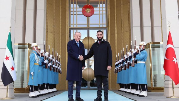 L'image montre deux hommes se serrant la main devant un bâtiment officiel. Ils sont entourés de gardes en uniforme bleu, tenant des fusils, et un grand symbole turc est visible derrière eux. La scène dégage une atmosphère solennelle.