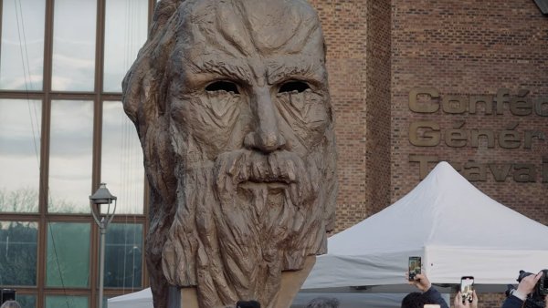 L'image montre une grande sculpture représentant un visage ancien, probablement celui d'une figure historique ou mythologique, avec des traits marqués et une barbe. Elle est entourée d'une foule de personnes qui semblent prendre des photos avec leurs téléphones. Il y a aussi des tentes et un bâtiment visible en arrière-plan, ce qui indique qu'il s'agit probablement d'un événement public ou d'une exposition. L'atmosphère semble être festive et attentive, centrée sur l'œuvre sculpturale.