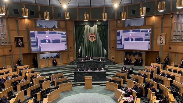 L'image montre l'intérieur d'une grande salle de réunion ou d'assemblée, probablement un parlement ou une assemblée législative. Au centre, une estrade est prévue pour les orateurs, et des écrans sont visibles sur les murs pour diffuser les interventions. On peut voir plusieurs membres assis sur des rangées en pente, organisés en sections. Les couleurs dominantes sont des tons de vert et de bois, créant une ambiance formelle. Des personnes sont également présentes debout, écoutant attentivement.