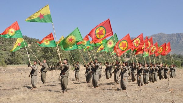 Des soldats en uniforme tiennent des drapeaux kurdes sur un terrain ouvert.