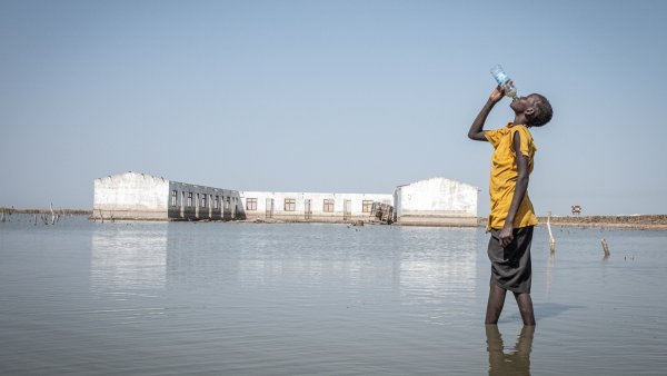 Un enfant, debout dans l'eau, boit de l'eau d'une bouteille, avec un bâtiment en arrière-plan.