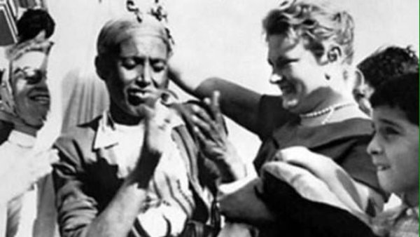 L'image montre un groupe de personnes dans une scène joyeuse et festive. Au centre, une femme souriante interagit avec un homme qui semble également être content. Ils sont entourés d'autres personnes, dont certaines affichent des expressions de bonheur. Les vêtements des personnes suggèrent une ambiance culturelle ou traditionnelle. L'atmosphère générale est celle d'une célébration ou d'un moment convivial.