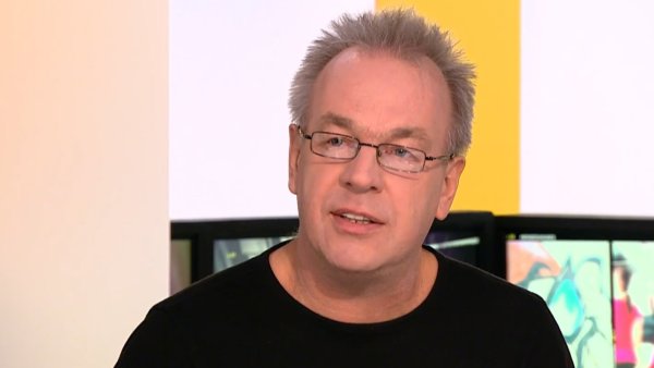 L'image montre un homme avec des cheveux gris et des lunettes. Il porte un t-shirt noir et semble en train de parler, avec un fond flou qui suggère un environnement de discussion ou d'interview.