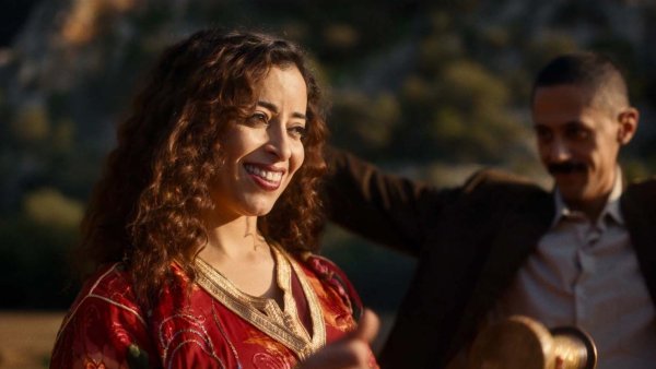 L'image montre une femme souriante avec des cheveux bouclés, portant une robe rouge ornée de motifs. Elle semble joyeuse et est entourée d'un environnement naturel. Un homme en arrière-plan, vêtu d'un costume, semble également profiter du moment. L'atmosphère générale est festive et conviviale.