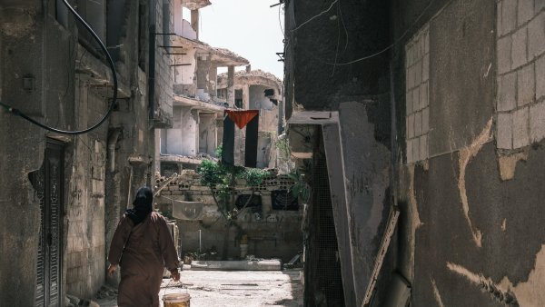 Une femme en manteau marron marche dans une ruelle étroite, bâtiments lézardés autour.