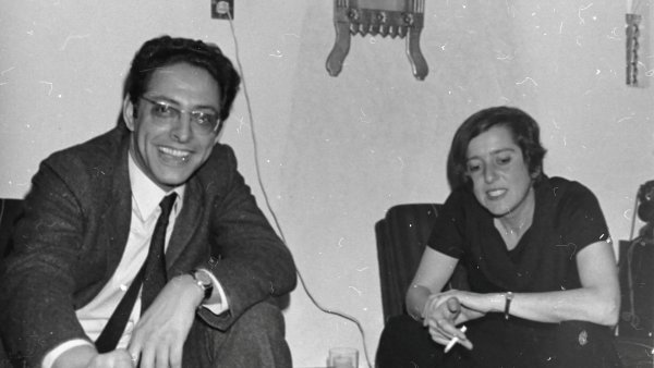 Deux personnes assises, souriantes, dans un intérieur sobre, une cigarette à la main.