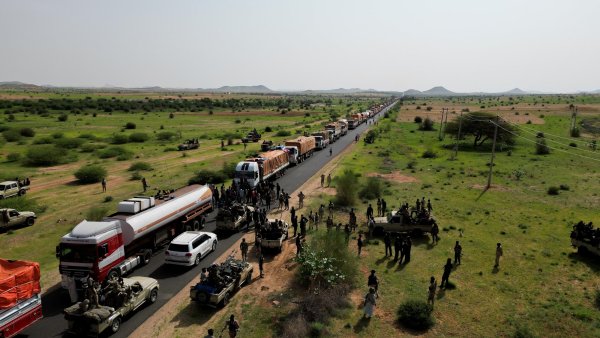 Convoi de camions sur une route, entouré de militaires et de verdure désertique.