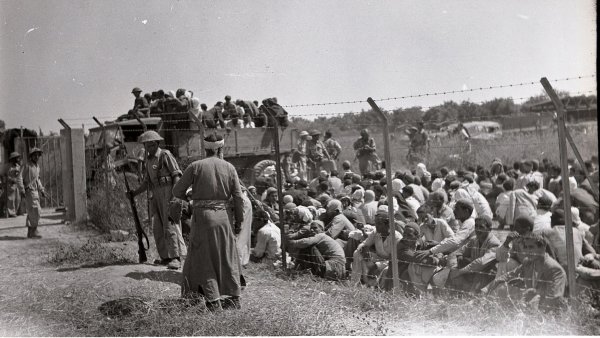 Des soldats surveillent un groupe de personnes assises derrière une barrière.