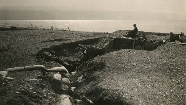 L'image représente un champ de bataille avec une tranchée. On peut voir un soldat assis sur les bords de la tranchée, qui semble surveiller les environs. Le terrain est accidenté et semé de sacs de sable, typiques des fortifications de la Première Guerre mondiale. À l'arrière-plan, on aperçoit une étendue d'eau qui pourrait être un lac ou une rivière. L'atmosphère générale est sombre et mélancolique, évoquant la dureté de la guerre.
