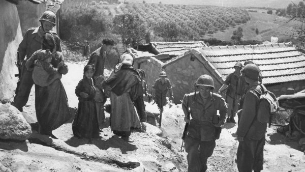 Soldats et civils en interaction dans un village, paysage verdoyant en arrière-plan.