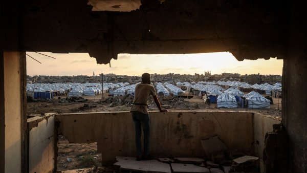 Un jeune observe un camp de tentes depuis une fenêtre endommagée au crépuscule.