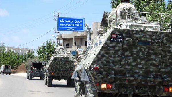 Des véhicules militaires avancent sur une route, près d'un panneau d'accueil.